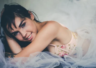 Gros plan d’une femme souriante portant un soutien-gorge floral rose allongée sur un tissu transparent blanc.